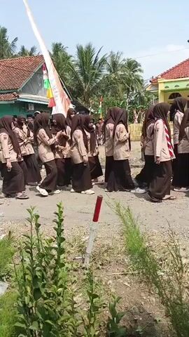 A group of young people in school uniforms, some wearing hijabs, are walking down a dirt road. Behind them, a group of young men in traditional clothing, some with face paint, are marching and playing instruments. The scene appears to be a parade or celebration. The environment is outdoors with houses and palm trees visible in the background. There are Indonesian flags decorating the area. The ground is dry and dusty, with some greenery growing at the bottom of the frame. A red and white striped pole is visible in the foreground.