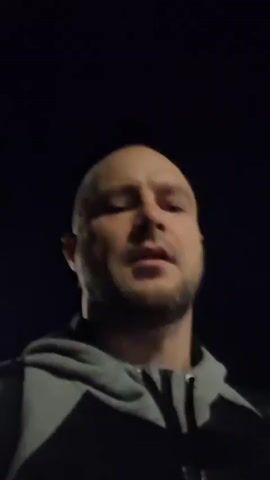 A man with a shaved head and a beard looks into the camera. He is wearing a grey hoodie with black accents. He is holding a baseball bat. The background is dark, suggesting it is nighttime. The man is speaking, but the audio is unclear.