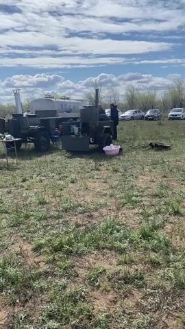 The video shows an outdoor gathering on a partially grassy field under a blue sky with scattered clouds. Several people are present, mainly gathered around cooking activities. A large pot sits atop a makeshift stove, emitting steam. Nearby, a white tent with green accents stands on the grass. Several cars are parked in the background, adding to the impression of an organized event in a natural setting.