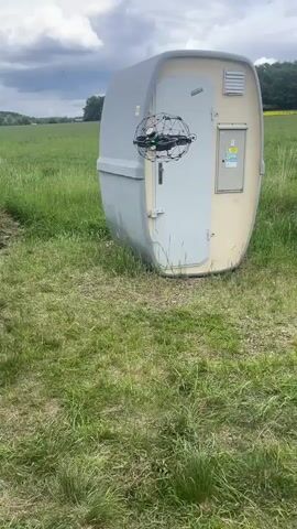 The video shows a small, black drone hovering near a large, egg-shaped structure in a grassy field. The structure, which is grey and beige, appears to be a type of utility enclosure or shed, with a door on one side. The drone has a protective cage around it and is positioned close to the structure's door. The field is green, and in the background, there are trees and an overcast sky. The drone is buzzing, and there are subtle sounds of nature. The camera angle shows the scene from a ground-level perspective.
