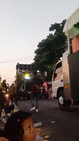 This is a vertical video clip, likely taken at dusk, showing a street celebration or parade. In the foreground, a young girl with dark hair and a pink clip looks up and to the right. The middle ground features a white truck with a stage set up on its bed, draped with white fabric and decorated with colorful lights. A banner on the side of the truck reads "XENIA MUSIC Makin Asyik." Several people are visible on and around the truck, including some standing on top of a large speaker stack behind it. The street is lined with trees and appears to be somewhat littered. The overall atmosphere is festive, with flashing lights and a sense of movement.