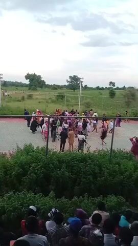 The video shows an outdoor gathering of people, likely spectators, watching an event on a court that appears to be for volleyball or basketball. In the foreground, a crowd of people is seated, some wearing hats and traditional clothing. The spectators are looking towards the court. The court itself has a net and is surrounded by a fence. Several individuals are on the court, some appearing to be playing the game and others observing. The environment is green with trees and grass visible in the background. The sky is cloudy. The camera is positioned at an angle, capturing both the crowd and the activity on the court.