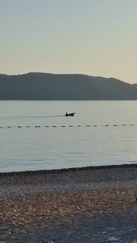 The scene is an outdoor shot of a beach and water, likely taken during a calm, possibly early morning, setting. In the distance, a small boat with a single person can be seen moving across the water. A line of buoys stretches out into the water, roughly parallel to the beach. The beach itself appears to be a mix of sand and small stones. In the background, there is a land formation, possibly a hill or a mountain. A portion of a wooden structure is visible on the right edge of the frame. The sky is a light blue color with a gentle gradient.