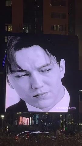 A large digital billboard displays a black and white portrait of a young man wearing a suit and tie. The man's face is shown in close-up, with different poses and expressions. The billboard is situated on the side of a building at night, with city lights and trees visible in the foreground. The text SHOQUANSUITS appears on the lower right corner of the billboard.