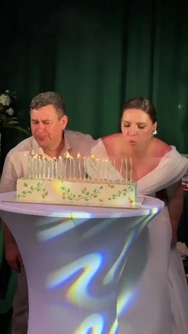 A man and a woman are positioned behind a table with a rectangular cake. The cake is decorated with green vine-like patterns and numerous unlit candles. The man, wearing a light-colored button-down shirt, and the woman, wearing a white off-the-shoulder dress, lean forward and blow out the candles on the cake. The woman has her hair pulled back and is wearing large earrings. The background consists of dark green curtains and some white floral arrangements. The table is covered with a white tablecloth that has a light projection of colorful, abstract shapes. The man and woman look at the cake as they blow, then look at each other, and finally at the camera.