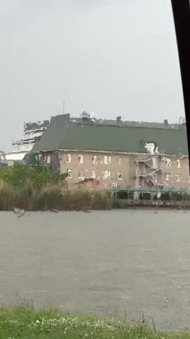 The video shows a building across a body of water. It's raining heavily, blurring the view. The building has a green roof and multiple windows. There are plants along the shoreline.