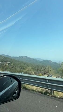 The video provides a first-person perspective from inside a moving car, driving through a scenic, mountainous landscape in Coajomulco, Mexico. The sky is clear and bright blue, indicating a sunny day. A grey guardrail runs along the side of the road, separating it from the hilly terrain. The landscape features rolling hills and distant mountains, covered with a mix of green and dry, sparse vegetation.

At one point, a white road sign with Spanish text becomes visible. The sign displays a yellow diamond with a black symbol of three humps (indicating speed bumps or an uneven road) and the text "Vibradores a 500 m," meaning "Speed bumps at 500 meters." Parts of the car's interior, including the side mirror and a glimpse of the dashboard, are visible in the foreground. Upbeat music with female vocals is playing in the background throughout the drive.