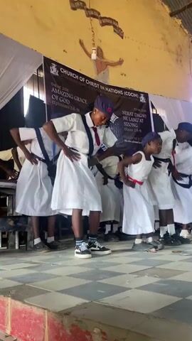 A wide shot shows a group of approximately 10 young people, mostly children, dancing on a small stage in what appears to be a church. The children are wearing white dresses or shirts with suspenders, and some have navy hats and red ties or sashes. They are moving in unison, clapping and stepping rhythmically, clearly performing a dance routine.

Behind them is a banner that reads "CHURCH OF CHRIST IN NATIONS (COCIN)" and has additional text about a theme of "AMAZING GRACE OF GOD." The room has a high ceiling, and there is a cross with a figure on the wall above the banner. The stage has a tiled surface and is painted with a rough, distressed look. The overall environment is modest, suggesting a simple church setting.