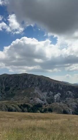 The video pans across a vast mountain landscape under a bright blue sky dotted with white clouds. The foreground features a field of dry, golden grass, while the middle ground transitions to rolling hills covered in sparse green vegetation and rocky outcrops. In the background, a large, rugged mountain range dominates the scene, with steep, craggy slopes and shadowed crevices. The overall impression is of a wild, natural, and somewhat desolate environment. There are no people, animals, or man-made objects visible in the footage.