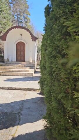 The camera pans left to reveal a small, white building with a brown door and a brown trimmed roof. There are steps leading up to the door. Green trees line the sides of the walkway. The sky is blue. The building has text written above the door.