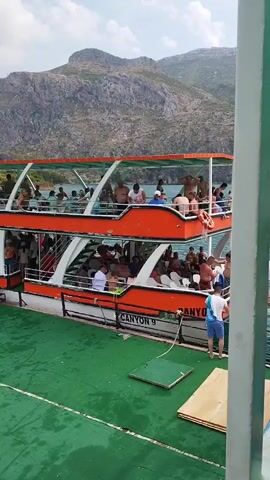A group of people are on a tour boat on a body of water. The boat has two levels with people on both. On the lower deck, several people are standing and sitting. A few people are near the edge of the boat, and one person is in the water. On the upper deck, people are standing and sitting. The boat is docked at a green-covered platform. In the background, there are mountains and trees. The weather appears to be sunny.