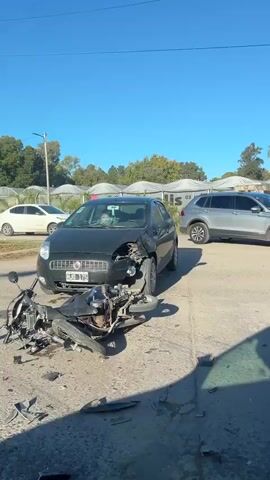 The video shows the aftermath of a car accident involving a car and a motorcycle. The black car has damage to the front, including a shattered headlight and a dented hood. The motorcycle is lying on the ground in pieces, with debris scattered around it. The car's license plate is visible, HJD-175. A man is visible sitting in the driver's seat of the car. Other vehicles are parked nearby. The accident appears to have occurred on a paved road with a sidewalk and trees in the background. The sky is clear and blue.