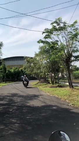 The video captures a sunlit day with a clear blue sky and some scattered clouds. A paved road curves through a grassy area dotted with trees. The road is shaded in places by overhanging branches. A person on a motorcycle is traveling away from the camera on the road. In the background, there is a large building with a sign that appears to read "TULANGAN". Power lines are visible strung across the sky. The overall environment suggests an outdoor setting, possibly a rural or suburban area.