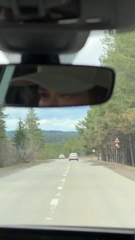 The video shows a person driving a car on a paved road lined with trees. The view is from the driver's seat, looking out through the windshield. In the distance, two cars are visible on the road ahead. The driver's eyes are visible in the rearview mirror, and they appear to be focused on the road. A navigation system is visible on the dashboard, displaying a map and distance to the destination. The sky is overcast with a mix of clouds.