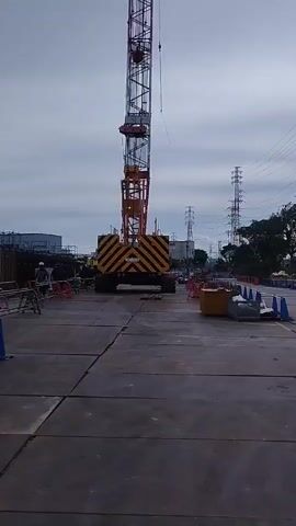 The video shows a construction site with a tall crane in the center. The crane has a yellow and black striped base and a tall, thin tower. There are several construction cones and barriers on the ground around the crane. In the background, there are power lines, trees, and buildings. The sky is cloudy.