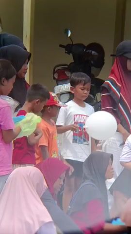 A group of children and adults are gathered outdoors. In the foreground, several children are standing and sitting. One boy in a white t-shirt holds a white balloon. Another child in a pink shirt holds a green plastic bag. Several adults are partially visible, some wearing hijabs. In the background, a motorcycle is parked. The scene appears to be a casual outdoor gathering.