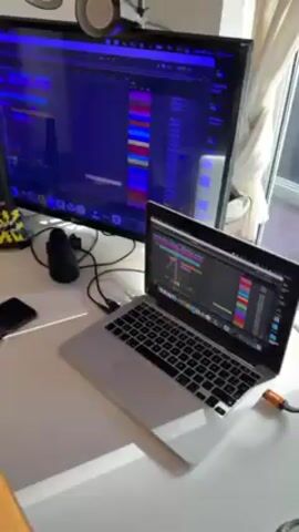 The video shows a laptop running Ableton Live music production software. The screen displays a timeline with colored audio and MIDI clips arranged in tracks. A larger monitor behind the laptop also shows Ableton Live. There are other peripherals on the white desk, including a microphone and cables. The room is well-lit.