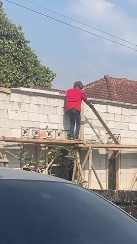 The video shows a person wearing a red shirt, dark pants, and a light-colored head covering, standing on a wooden platform. The person appears to be a construction worker, actively engaged in building a wall made of white bricks. They are positioned on the platform, facing away from the camera, and seem to be working with a plank of wood. To the left, a partially visible red and white flag is present. The background consists of a tiled roof, greenery, and a bright blue sky. The overall scene suggests an outdoor construction site, likely a residential area.
