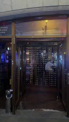 The video shows the exterior of a building with an open doorway. The doorway leads into a dimly lit interior decorated with a wall of sparkling, diamond-shaped ornaments. Inside, at least two people are visible. One person, wearing a white shirt, is seated at a table on the right. Another person, in a blue shirt, is partially visible behind a patterned curtain to the left. The entrance area is tiled, and a silver trash can is located to the left of the doorway. A sign above the entrance is partially visible and reads "CONDICIONED ENTRANCE FLOORS." The lighting is predominantly purple and yellow, with blue and pink accents from the decorations.