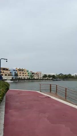 This media file depicts a scenic waterfront view, likely in Higüey, Dominican Republic. The scene is dominated by a body of water, possibly a canal or bay, with a vibrantly colored boardwalk or path in the foreground. This path is a striking reddish-pink, contrasting with the muted tones of the sky and water.

Across the water, a row of buildings is visible, featuring a variety of pastel colors, including light yellow, orange, mint green, and white. These buildings suggest a resort or residential area with a Mediterranean or Caribbean architectural style, characterized by their stucco exteriors and tiled roofs. Some of the buildings are multi-story, with visible balconies and windows.

Further in the distance, more modern white buildings are present, along with lush green trees and palm fronds, indicative of a tropical climate. A small boat or vessel can be seen in the water, suggesting recreational or possibly transport activities.

The sky is overcast and a uniform grey, hinting at cloudy weather, possibly a light rain, as the boardwalk appears slightly damp in places. The overall mood is calm and serene, with the soft, diffused light of an overcast day. There are no people clearly visible in the frame, and no discernible text is present in the media. The perspective is from ground level, looking out over the water towards the buildings. The presence of a wooden railing on the right side of the frame indicates a pier or promenade.