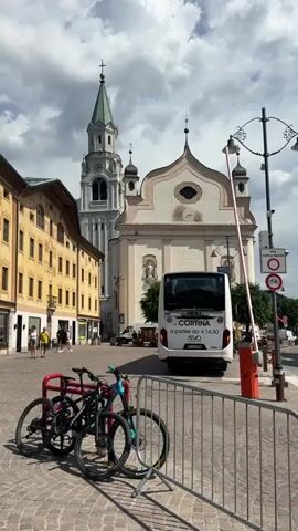 The video opens with a view of a European town square, likely Cortina d'Ampezzo, Italy, given the text on the bus. In the foreground, two bicycles are parked at a red rack, secured by a metal fence. The bikes appear to be mountain bikes. Beyond the fence, a white tour bus with "CORTINA" and "a partire da 14.80" visible on its rear is stopped, with its back to the camera. To the left of the bus is a building with classical architecture, and behind it, a tall church tower with a green spire dominates the skyline. The church itself has a pale facade with a domed roof.

The scene is set on a cobblestone street, with buildings lining one side of the square. Several people are visible walking in the background, dressed in casual summer attire. The time of day appears to be daytime, possibly late morning or early afternoon, indicated by the quality of light and the presence of clouds in the sky. The weather seems to be mixed, with sunshine breaking through partly cloudy skies.

Notable details include street signs, one of which appears to indicate a pedestrian zone and prohibits bicycles. The overall atmosphere is peaceful and scenic, suggesting a typical day in a picturesque Italian town. The emotion conveyed is one of everyday life in a tourist destination. The text on the bus and the signage suggests transportation and local regulations are relevant aspects of the scene.