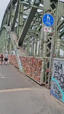 A white high-speed train with red accents travels across a green metal bridge. The bridge has a pedestrian and bicycle path on the left, lined with numerous colorful love locks attached to the railing. Several people are walking on the path. A blue circular sign depicting a pedestrian and a bicycle is mounted on a pole. The bridge structure is visible, with its intricate metalwork. Graffiti is present on a metal box or panel on the right side of the image.