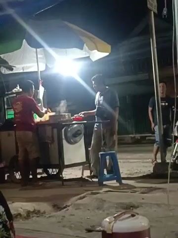 A man in a red shirt and tan shorts stands behind a food cart, seemingly preparing food. Another man, wearing a blue shirt and tan pants, stands in front of the cart, holding onto a horizontal bar. A blue stool is placed near the second man. In the background, a white car passes by. To the right, another man in a black t-shirt and shorts stands near a motorcycle. The scene appears to be an outdoor market or street food vendor at night, illuminated by a bright light from the food cart's umbrella. There is steam rising from the food cart.