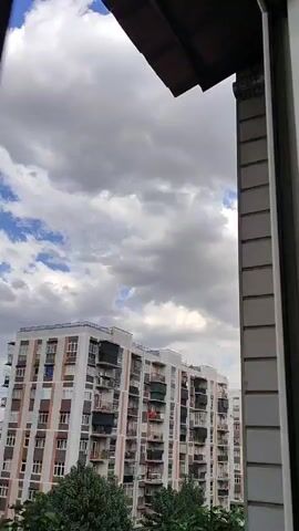 The video shows an upward view from a window looking at a cloudy sky and a residential building. The building is tall with multiple floors and many windows. There are trees in the foreground partially obscuring the lower part of the building. The clouds are dense and appear to be moving, suggesting a dynamic sky. The right side of the frame is dominated by a portion of another building or structure, possibly part of the same window frame, with vertical siding. The overall impression is a view of an urban environment on a cloudy day.
