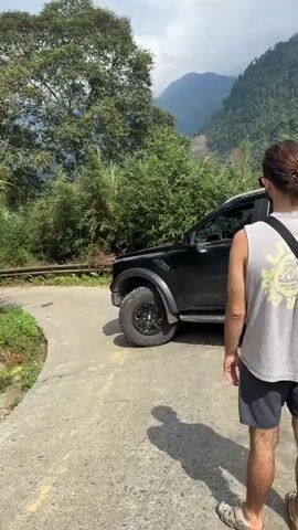 A black Ford Ranger truck cautiously navigates a narrow concrete bridge. The driver is visible through the side window, maneuvering the vehicle. A person in a gray tank top and gray shorts stands on the side of the bridge, casting a shadow on the concrete. The bridge is bordered by greenery and a small drop on one side. Lush foliage and mountains form the backdrop.
