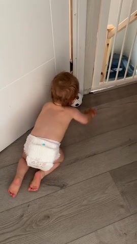 The video shows a baby, seen from behind and from above, wearing a diaper. The baby has light brown hair. The baby is on a wooden floor, near a white door or wall. The setting appears to be indoors, possibly a home.

In the first few seconds, the baby is crawling or kneeling, looking towards an opening to the right, which seems to be a gate leading to stairs. A small black and white soccer ball is present.

The baby then interacts with the soccer ball. The ball bounces off the wall or door. The baby attempts to hit the ball with their hands. The baby is playing with the ball on the floor.

The time of day and weather are not discernible from the video. There is no visible text in the video. The location cues suggest it is indoors, and the context provided states Leeds, United Kingdom. The baby's actions indicate playful engagement with the toy.