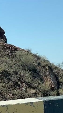 The video shows a roadside view from a moving vehicle. The scene is dominated by rocky hills and a clear blue sky. In the foreground, a yellow and black guardrail is visible. The landscape transitions from rugged, rocky terrain with sparse vegetation to a wider vista revealing a body of turquoise water and a town or city in the distance. The water appears to be a reservoir or lake, with several boats visible on its surface. The town features buildings and some signs of human settlement. The overall impression is of a scenic drive through a mountainous and water-rich region.
