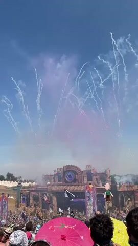 The video shows a wide shot of an outdoor music festival. In the background, a large, elaborate stage is adorned with a circular eye motif and intricate structures. Fireworks are exploding in the sky above the stage, casting a smoky haze and colorful trails. A crowd of people is gathered in front of the stage, many with their arms raised in the air. In the foreground, several people are visible, including a person holding a bright pink umbrella. The sky is blue with some scattered clouds. The overall atmosphere appears to be festive and energetic.
