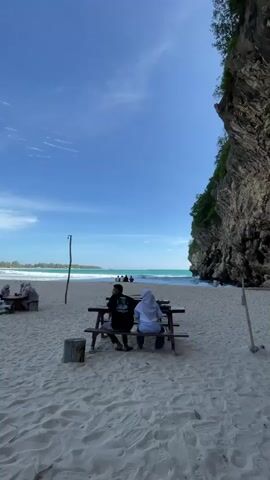 The video shows a serene beach scene with a bright blue sky and white sand. In the foreground, two individuals are seated at a wooden picnic table, facing the ocean. The person on the left is wearing a black shirt, and the person on the right is wearing a white headscarf and a white top. Behind them, several other people are scattered across the beach, some sitting and others standing. The ocean is a vibrant turquoise color, with waves gently rolling towards the shore. To the right of the frame, a large, rocky cliff covered in lush greenery rises dramatically from the beach. The overall atmosphere is one of tranquility and relaxation.