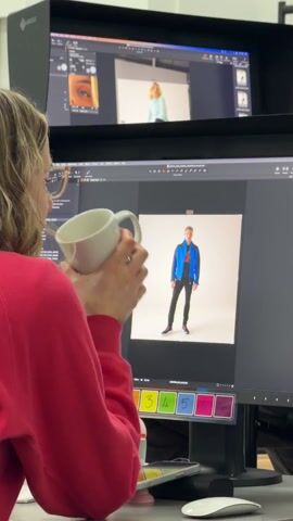 The video shows a woman in a red sweatshirt taking a sip from a white mug in front of a computer screen displaying a photo shoot. The video cuts to a shot of the woman's back as she walks over to two women sitting on the floor in a white studio space. One woman is wearing a light green shirt and the other is wearing a black shirt. A brown jacket is draped over a chair in front of them.