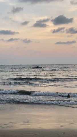 Here is a description of the video:

The video shows a beach scene at dusk. The sky is a blend of pinks and grays with scattered clouds. In the distance, the ocean horizon is visible, with a boat moving across the water. Closer to the shore, waves are gently lapping at the sandy beach. Several people are walking along the shoreline, including a woman in a white hat and a patterned top, and two women in swimsuits. 

The scene then shifts to show people standing and taking photos of parasailers in the sky. There is a man in a striped shirt standing near a banana boat on the beach. The parasails are colorful and high above the ocean. Other beachgoers are in the water in the background.
