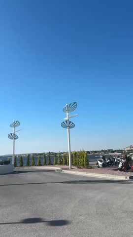 The video opens with a view of an empty paved area leading towards a waterfront. Two distinctive white sculptures, one resembling a large heart with an opening and the other a cluster of leaf-like shapes on poles, are visible. Beyond these, a calm body of water stretches towards a distant tree-lined horizon. The sky is a clear, bright blue. As the video progresses, the perspective shifts, offering a wider panorama of the waterfront. More of the stylized sculptures are seen, along with neat rows of green trees lining the shore. The sun is bright, casting long shadows. The video then pans across a grassy area with more of the unique tree-shaped sculptures, and a few people are seen walking along a paved path. The scene appears to be a public park or a scenic waterfront promenade. The presence of metal barriers suggests a controlled access area. The overall impression is one of a modern, well-maintained outdoor space bathed in sunlight.