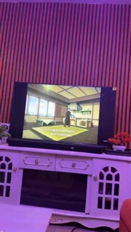 A flat-screen television displays a scene from an animated show featuring a character speaking the line, "Things are looking up, uncle." The TV is positioned on a white entertainment center with decorative accents. The room has a red and blue striped wall in the background. Additional decorative items, including plants, are present on the entertainment center. The lighting in the room appears to have a purple hue.