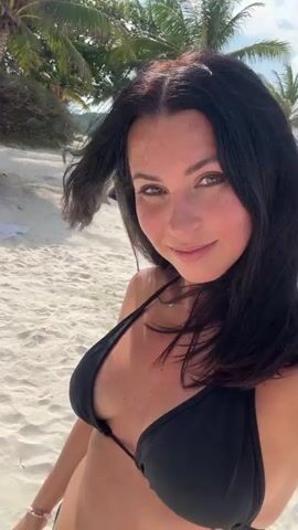 The video shows a woman in a black bikini standing on a white sandy beach. She is facing the camera and smiling. She has long, dark hair that is blowing in the wind. There are palm trees in the background.