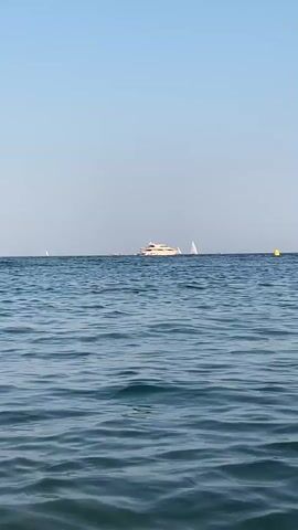 The video shows a wide shot of a calm ocean under a clear blue sky. In the distance, a large white yacht is visible on the horizon, with several sailboats around it. There is also a yellow buoy in the water to the right of the yacht. The foreground of the video is dominated by the rippling surface of the ocean. The camera is static, and no people are clearly visible in the frame.