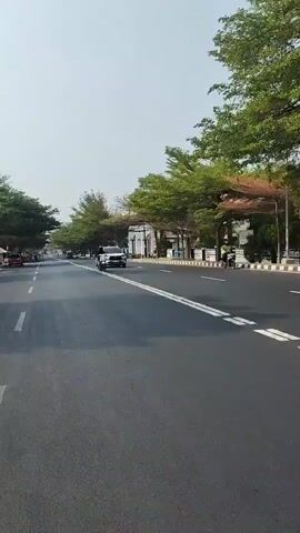 The video shows a street scene from the perspective of someone riding a motorcycle. The road is paved with asphalt and has white lane markings. On either side of the road are trees with green and some reddish-brown leaves, indicating it may be autumn or a similar season. There are buildings and some structures along the sides of the road, including what appears to be a small stand or kiosk on the left. Several vehicles are visible: a white car is in the right lane, a motorcycle with a rider is ahead of the camera in the right lane, and a red car is parked on the left side of the road. Further down the road, more vehicles, including a yellow truck, can be seen. The sky is bright and appears to be clear. The overall impression is of a well-maintained urban or suburban street on a clear day.