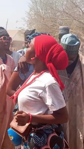 A group of people are gathered outdoors in daylight. Some are dancing, and many are wearing traditional clothing. A woman in a white shirt, red headscarf, and patterned skirt is prominently featured and moves through the crowd. The scene appears lively, with people interacting and moving around. The environment includes bare trees and dusty air.