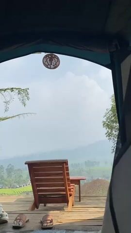 The video opens with a view from inside a tent, looking out towards a scenic landscape. In the foreground, two pairs of sandals are visible on a wooden deck, along with a white shoe and a pair of white platform shoes. A wooden chair and a small table are positioned on the deck, facing the view. Beyond the deck, a vast expanse of rolling hills covered in green foliage stretches into the distance. The sky is partly cloudy and a wicker light fixture hangs from the tent ceiling. The scene appears to be a campsite with a picturesque view. The video pans slightly to reveal more of the tent's interior, where a small wooden table holds various items, including bottles, cans, and what appears to be a small cake or pastry. The tent entrance frames the outdoor scenery.