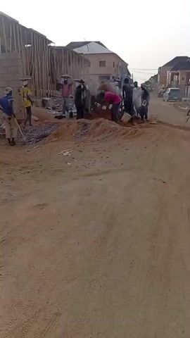 The video shows a construction site on a dirt road in an urban area. About a dozen people are working around a cement mixer and piles of gravel. Some carry materials on their heads. A bicycle passes by.  There are several completed buildings nearby.
