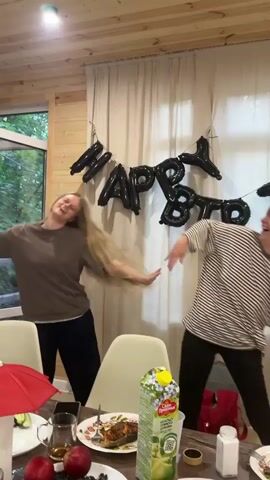 Two women are dancing in front of a "HAPPY BIRTHDAY" banner made of black balloons. The woman on the left has long blonde hair and is wearing a brown t-shirt and black pants. The woman on the right has short brown hair and is wearing a striped t-shirt and black pants. They are in a room with wood-paneled walls and a window with a view of trees. A table with food and drinks is in the foreground. The woman on the right is laughing and appears to be enjoying herself. The woman on the left is also smiling and dancing energetically.