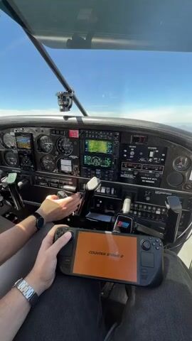 The video shows a person holding a black handheld gaming device displaying the game Counter-Strike 2 in front of an airplane cockpit dashboard. The person's hands are visible, one wearing a watch, holding the device. The other hand is near the aircraft controls. The cockpit instruments and windshield are also visible, with a clear sky and land visible outside.