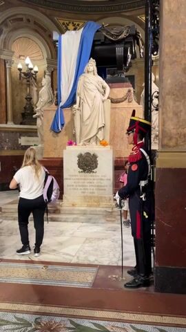 The video shows the interior of a building, specifically a mausoleum or crypt, with a large white marble statue as the focal point. The statue, draped partially with an Argentinian flag, stands atop a marble base with inscribed text and adorned with flowers. The text mentions "TRIUNFO EN SAN LORENZO-1813" and references independence and emancipation in various South American countries.

Several people are present in the scene. A young woman with long blonde hair and a purple backpack walks toward the statue. To the side, a uniformed guard stands at attention, holding a saber. Other individuals, including a child in a pink shirt, are seen in the background, observing the monument.

The environment is characterized by marble floors, ornate pillars, and arched ceilings. Additional busts are positioned around the main statue. The overall atmosphere suggests a place of historical significance and reverence.