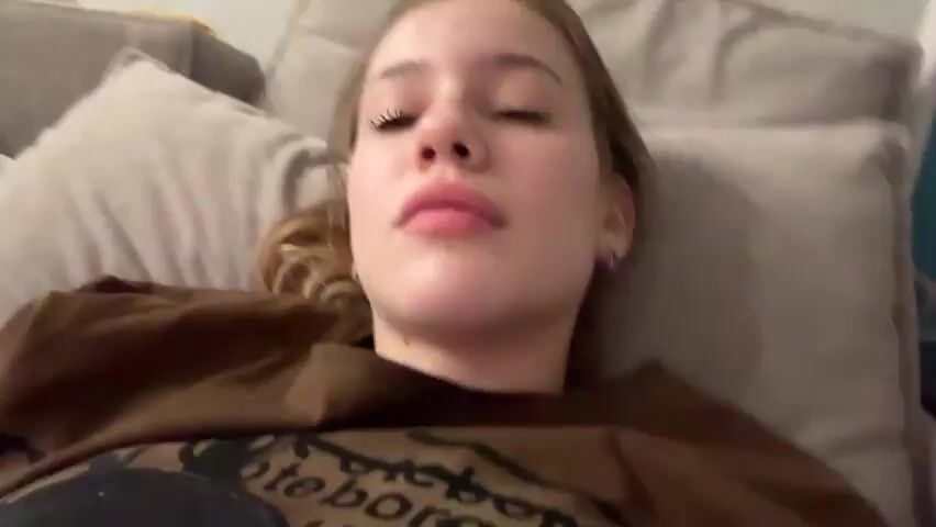 A young woman lies on a bed, looking at the camera. She is wearing a brown t-shirt with a graphic print. Her hair is light brown and pulled back. She is looking directly at the camera and appears to be speaking. The background is a pillow and a blanket.