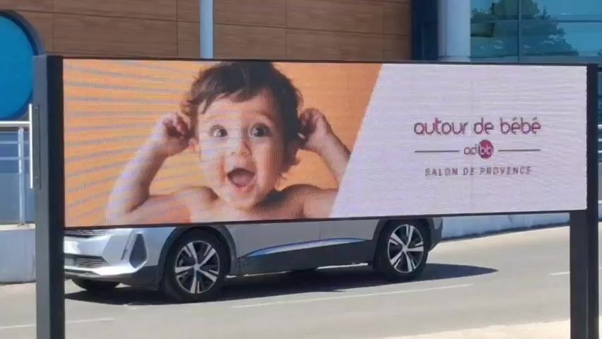 A digital billboard displays an advertisement for "Autour de Bébé Salon de Provence." The left side of the billboard features a close-up image of a laughing baby wearing pink headphones. The right side of the billboard has text that includes the company logo, the name of the event, and a list of products and services such as "LISTE DE NAISSANCE - ARTICLES DE PUERICULTURE - DEMONSTRATION ET CONSEILS PRODUITS - HYGIENE." Below this, the address "279 VIEUX CHEMIN D'ISTRES A SALON-DE-PROVENCE" is shown, with a parenthetical note indicating its location behind an Intermarché, formerly Géant Casino. The billboard is mounted on two black posts and is situated outdoors, likely roadside, with a street and a car visible in the background.