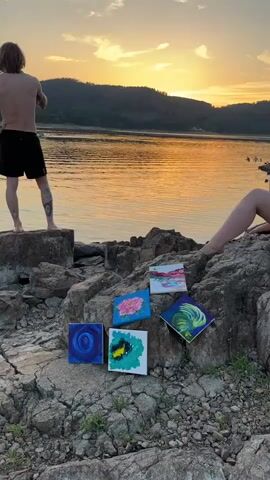 At 00:00, a person with long, blond hair, bare-chested and wearing black shorts, stands on a large rock overlooking a body of water at sunset. Their left leg is extended, and they appear to be gesturing with their right hand. In the foreground, on a rocky shoreline, are five small, square paintings. From left to right, the paintings depict a blue spiral, a pink flower, a yellow and black abstract design, a white and pink abstract design, and a green and blue feather-like design. The sun is setting behind rolling hills, casting an orange and yellow glow across the water. Another person, partially visible from the legs down, is sitting on rocks to the right. At 00:01, the camera slightly zooms in on the paintings. At 00:02, the camera pans slightly to the right. At 00:03, the camera continues to pan right, showing more of the shoreline and the water. At 00:04, the camera is focused on the paintings and the person's legs, with the sunset in the background.