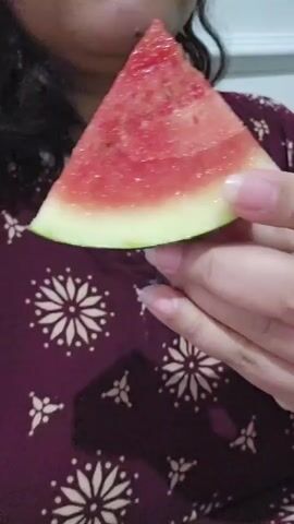 The video shows a close-up of a person holding a slice of watermelon to their mouth. The watermelon is a wedge shape with green rind and pink flesh. The person's mouth is open and they are taking a bite of the watermelon. The person is wearing a dark red shirt with a white floral pattern. The background is out of focus but appears to be indoors.